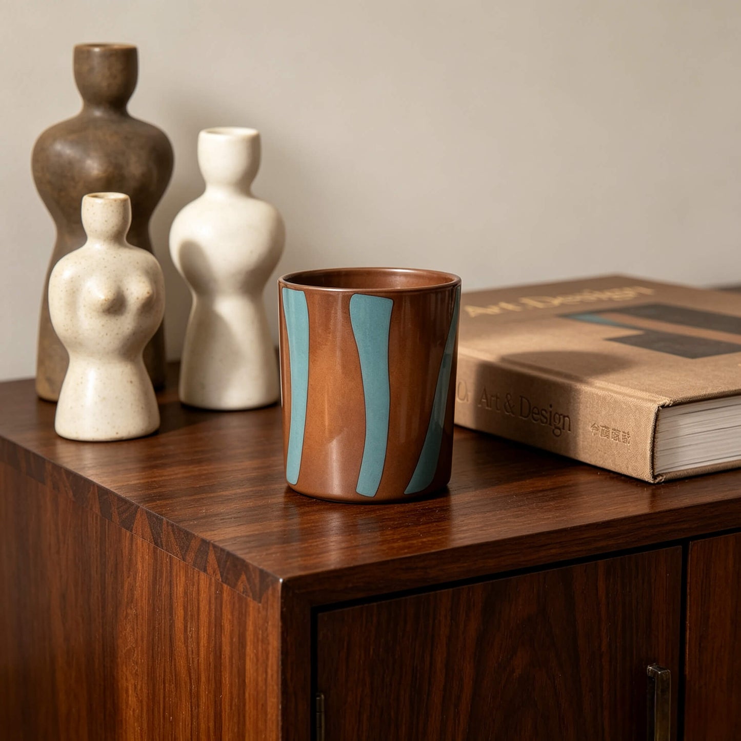 Empty terracotta and green striped candle vessel on a mid century modern wood cabinet with artistic statues