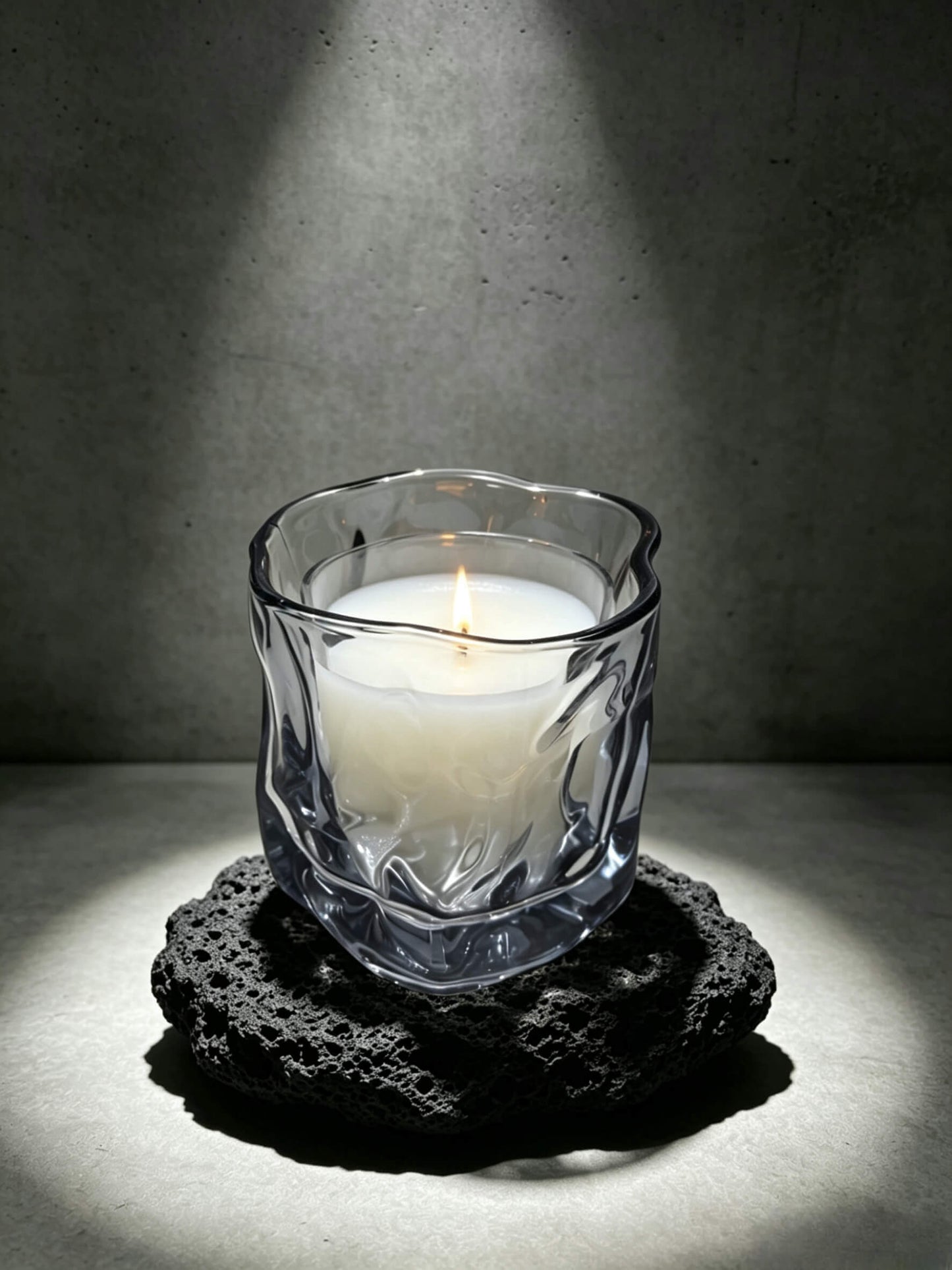 Lit smoky quartz wabi sabi folded candle vessel resting on a dark brutalist lava stone