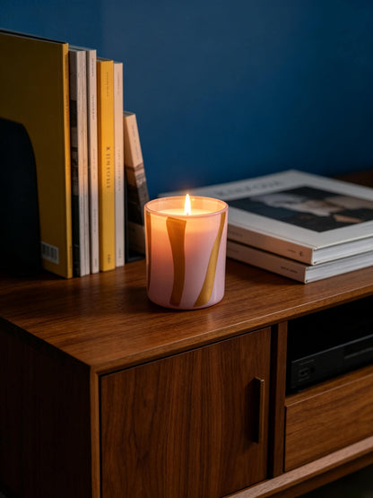 Lit pink and yellow striped glass candle jar casting a warm glow on a walnut credenza next to books for a moody cozy aesthetic