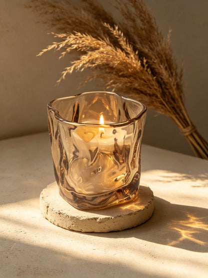 Lit amber wabi sabi folded candle vessel glowing on a stone coaster in sunlight with dried pampas grass