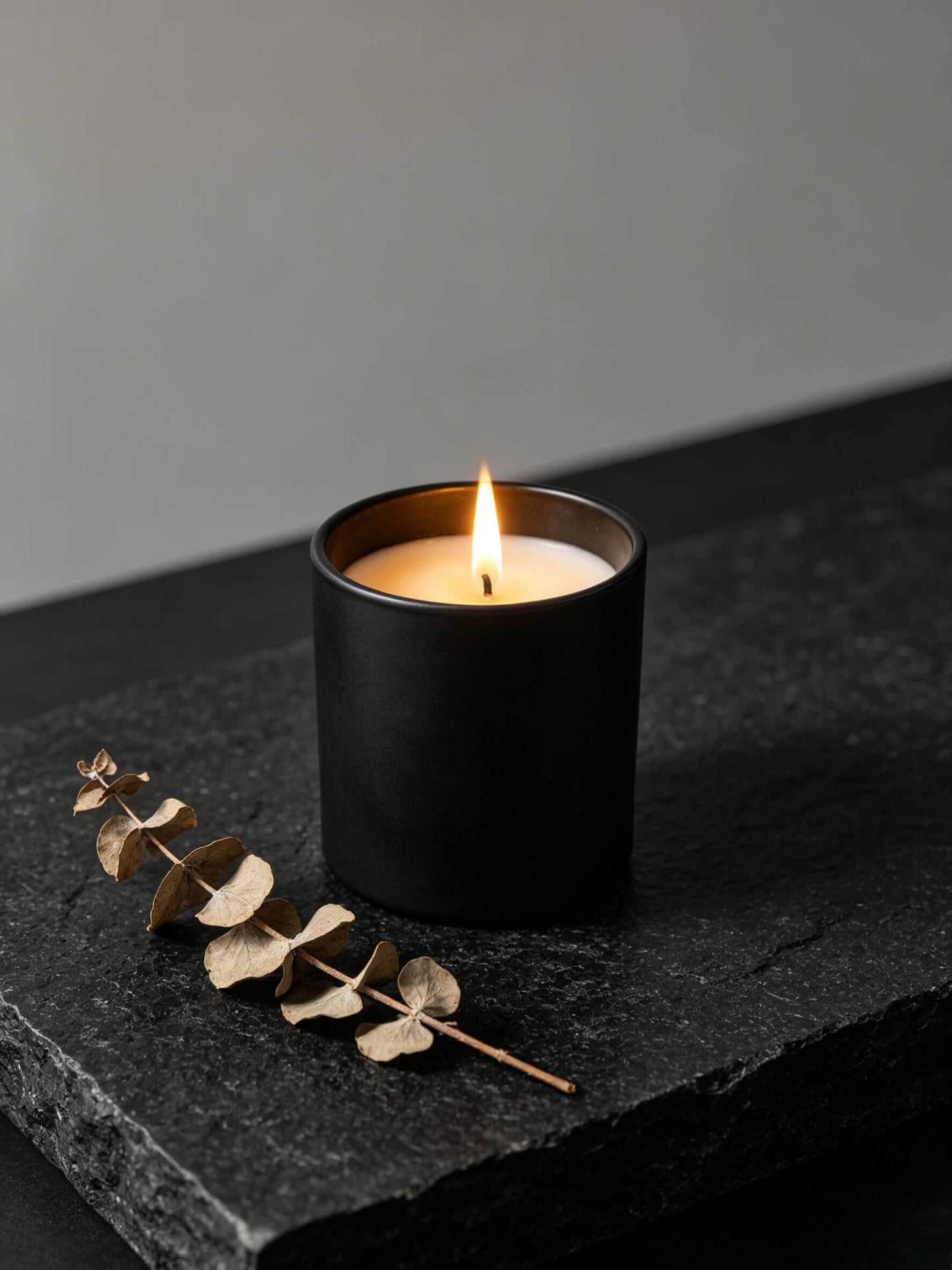 Lit matte black candle vessel styled with dark stones and dry branch for moody holistic wellness decor